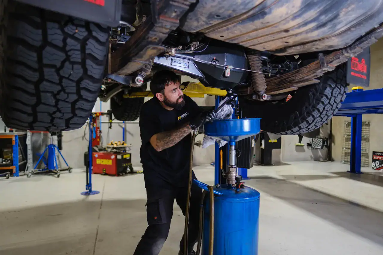 An M4C technician working under a 4WD on the hoist in the Canning Vale workshop, rear leaf springs and shackles overhead